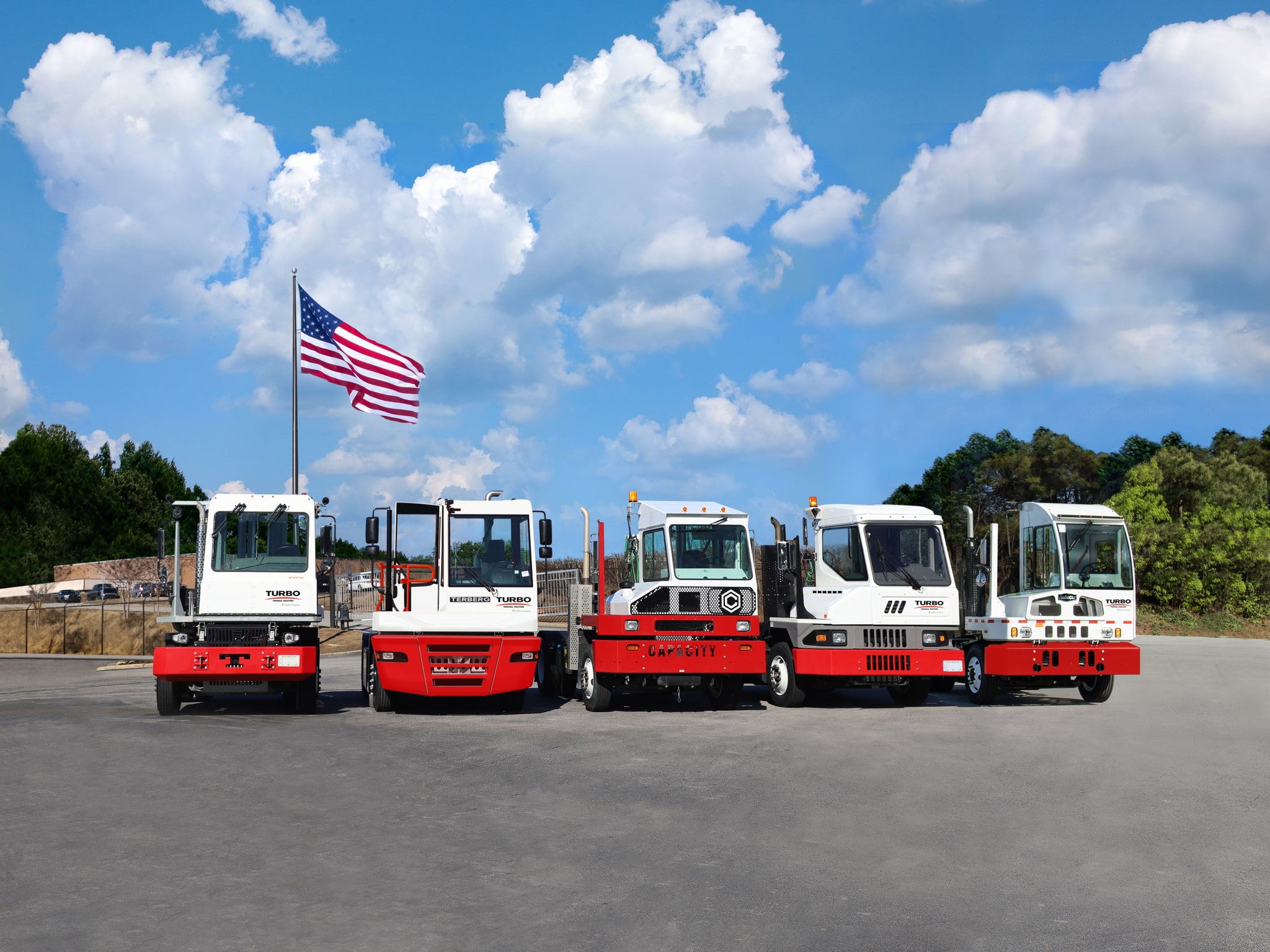 5 Turbo Terminal Tractor Spotter Trucks for sale lined up on the concrete in front of the American flag and pretty blue sky.